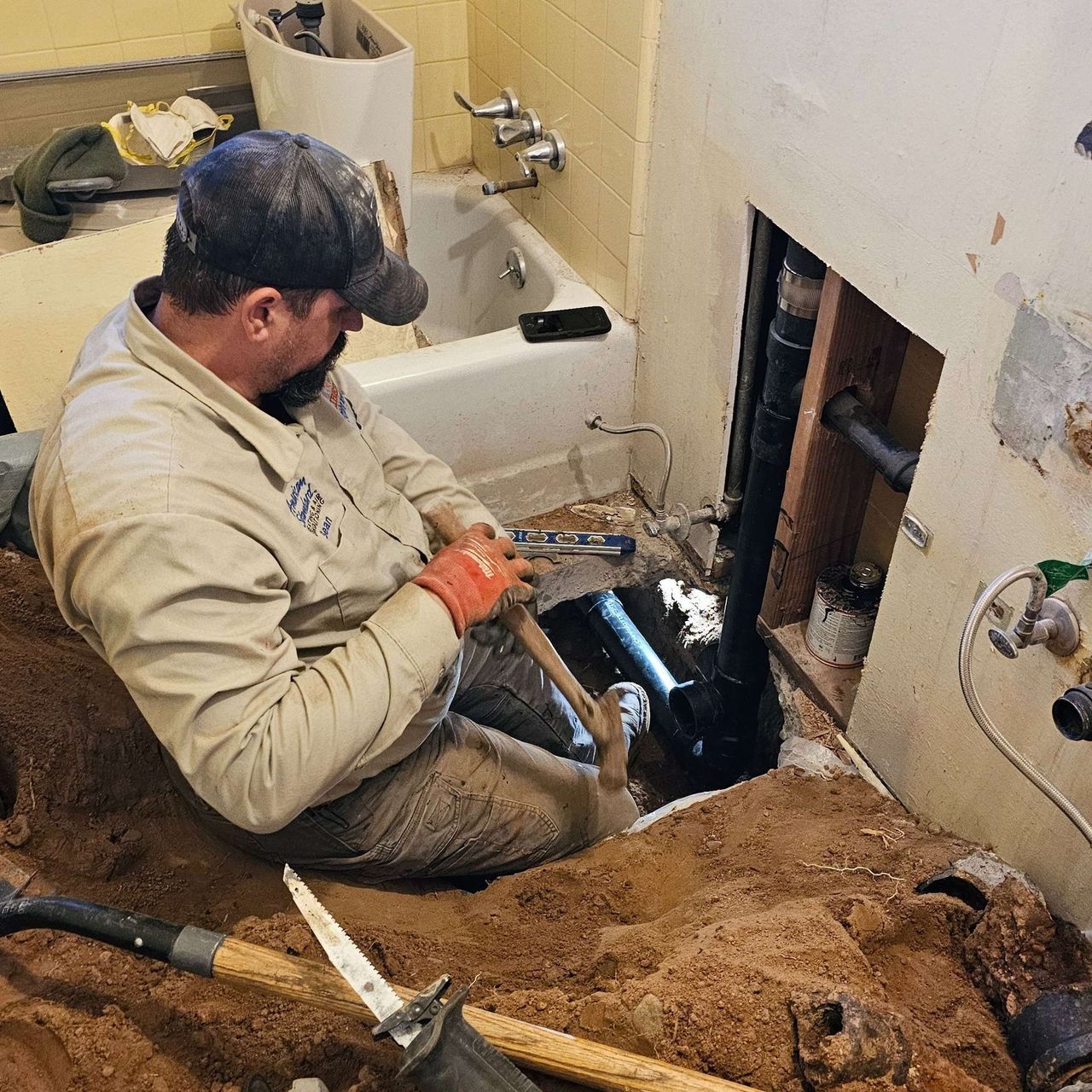 Plumber working on bathroom pipes in a hole. Tan uniform, red gloves, setting is a bathroom.