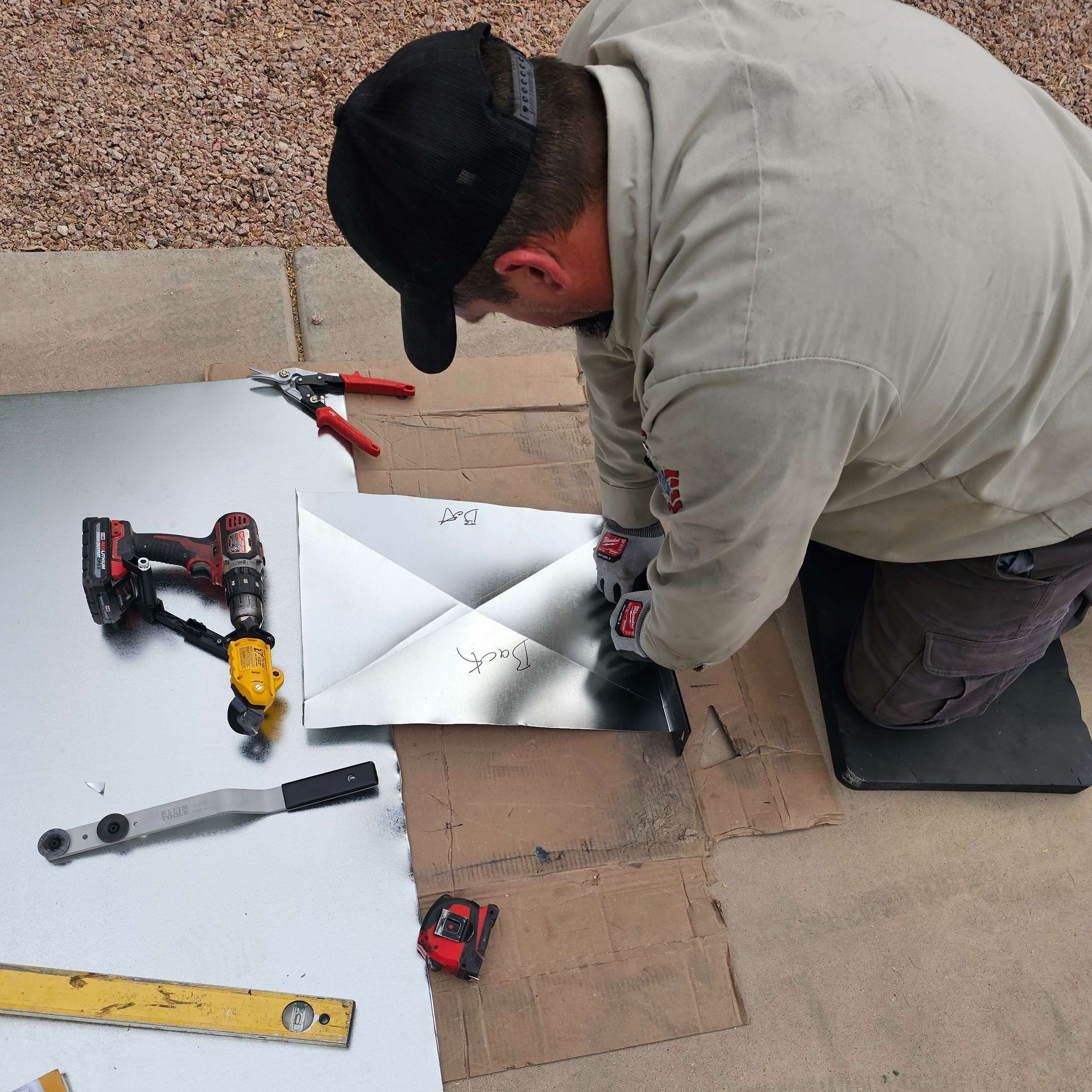 Man kneels, working with sheet metal. Tools like drill, pliers, level, and tape measure surround him outdoors.