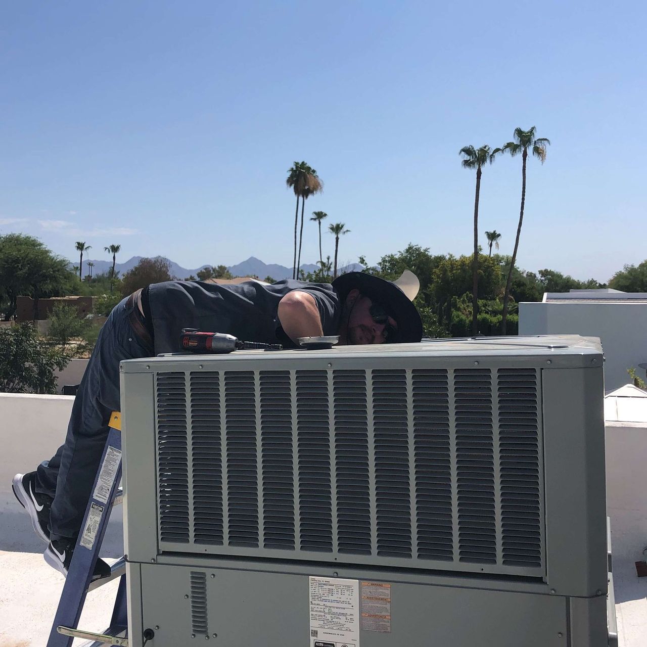 Man on a ladder repairing AC unit on a roof; sunny day, palm trees in the background.