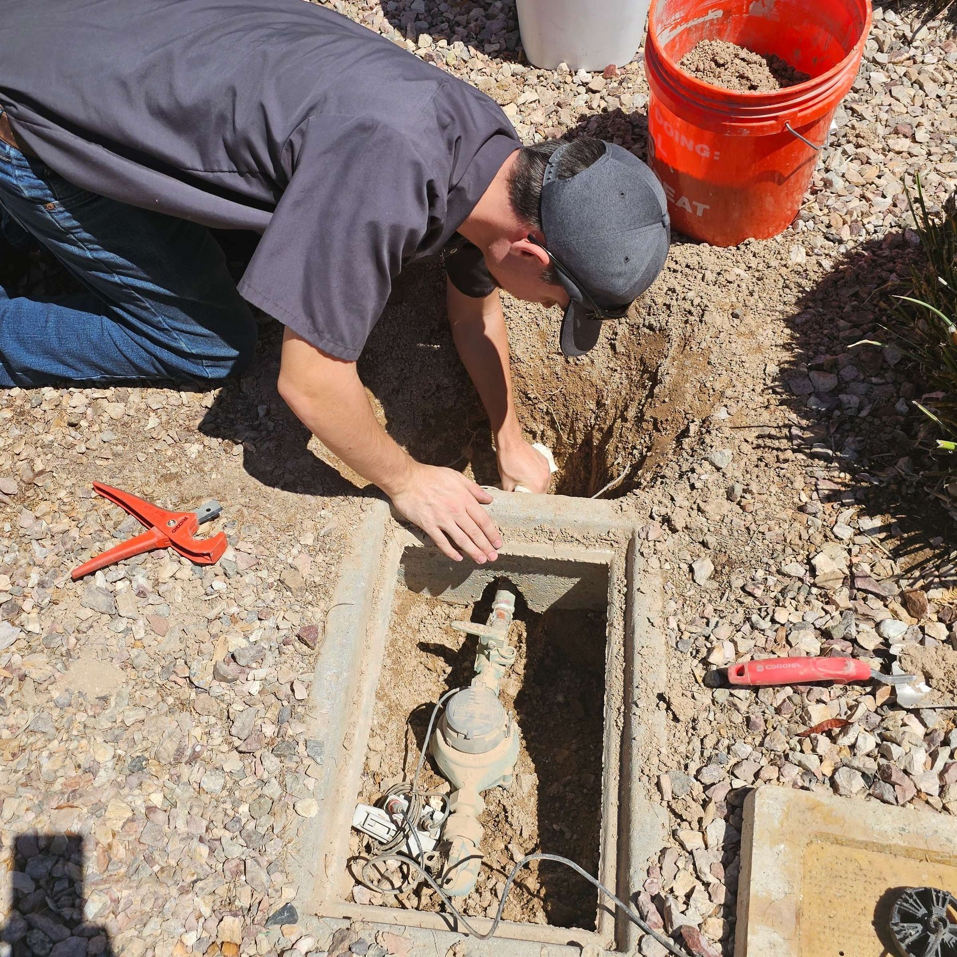 Man kneeling, working on plumbing in a concrete box in a yard with reddish gravel.