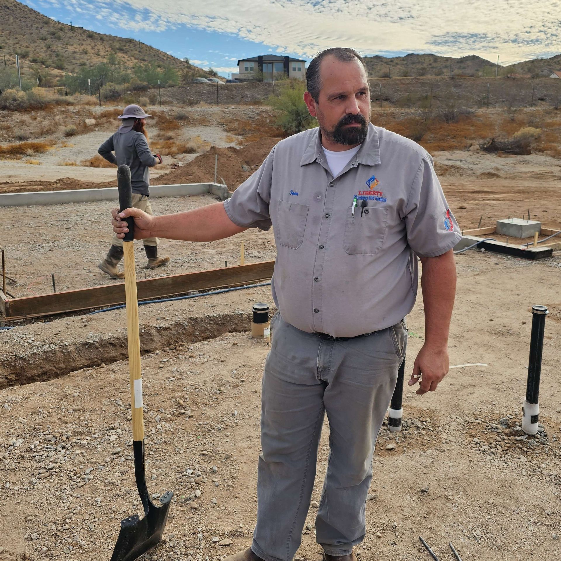 Construction worker with a shovel on a worksite, looking to the side. Another worker in the background. Sunny day.