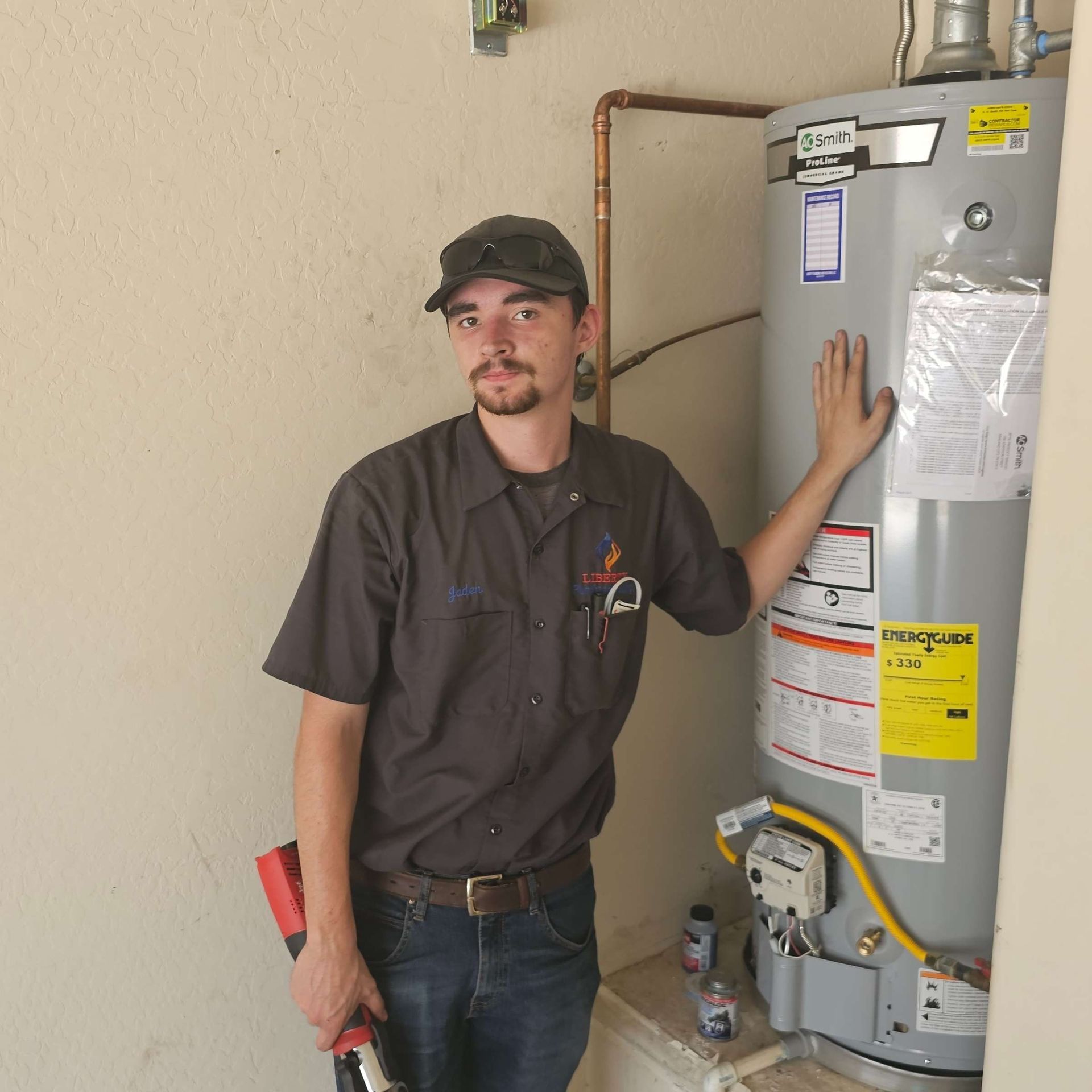 A plumber in a dark shirt and cap leans on a hot water heater, holding pliers.