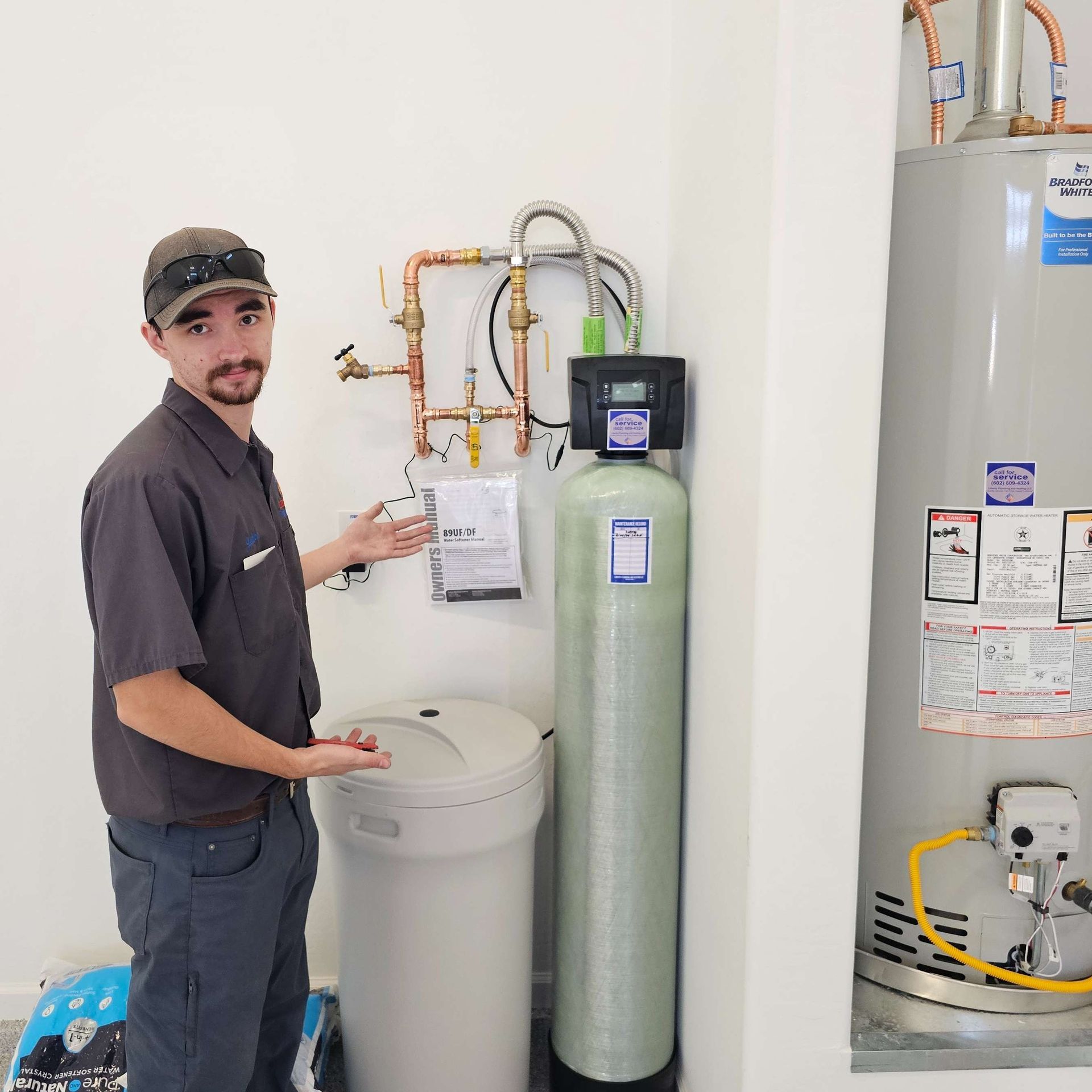 Man in work clothes points to water softener system and water heater in a utility room.