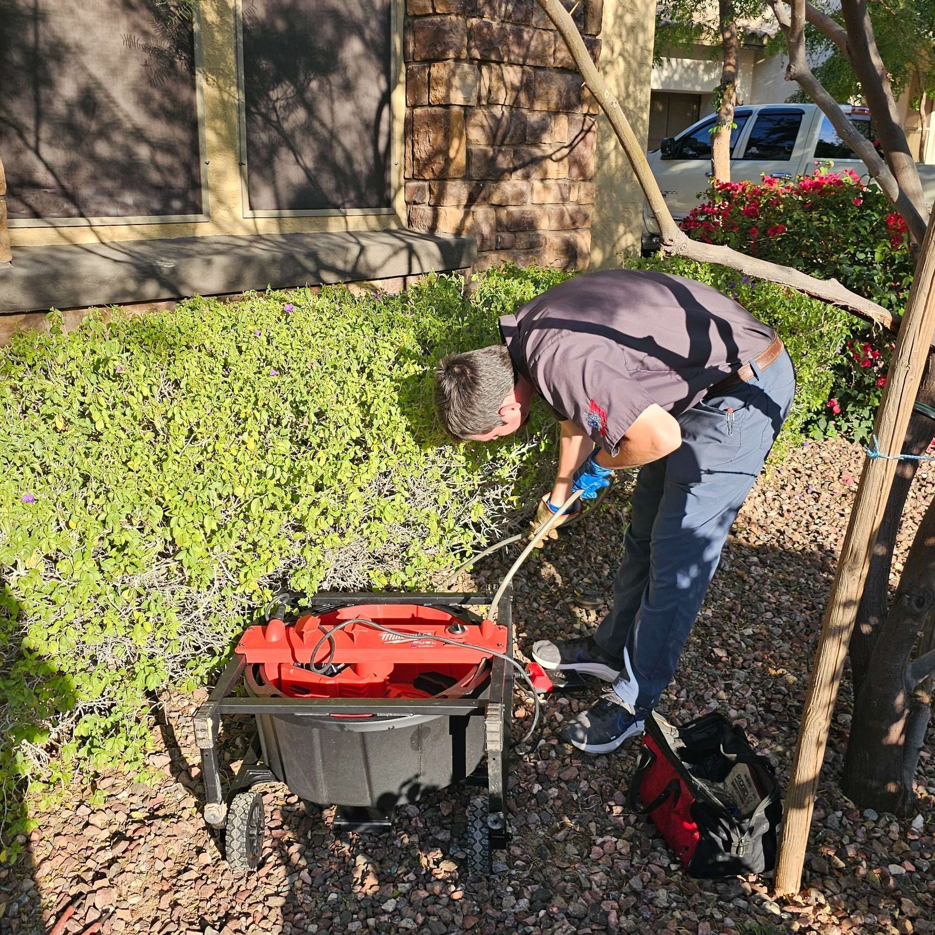 A man sprays a bush with a red sprayer. He's next to a house, wearing a dark shirt and jeans.