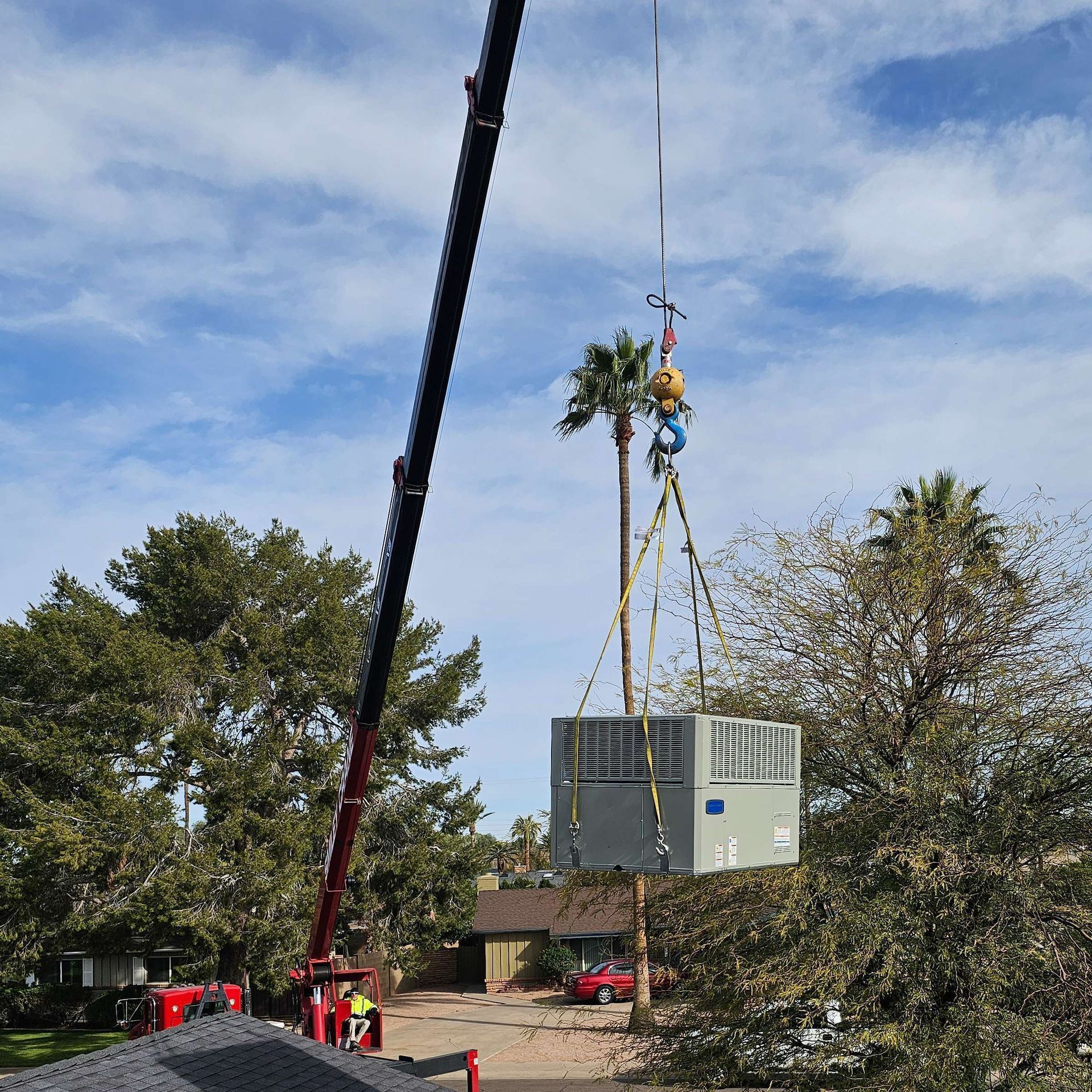 Crane lifting a large HVAC unit over a residential area with palm trees and cloudy sky.