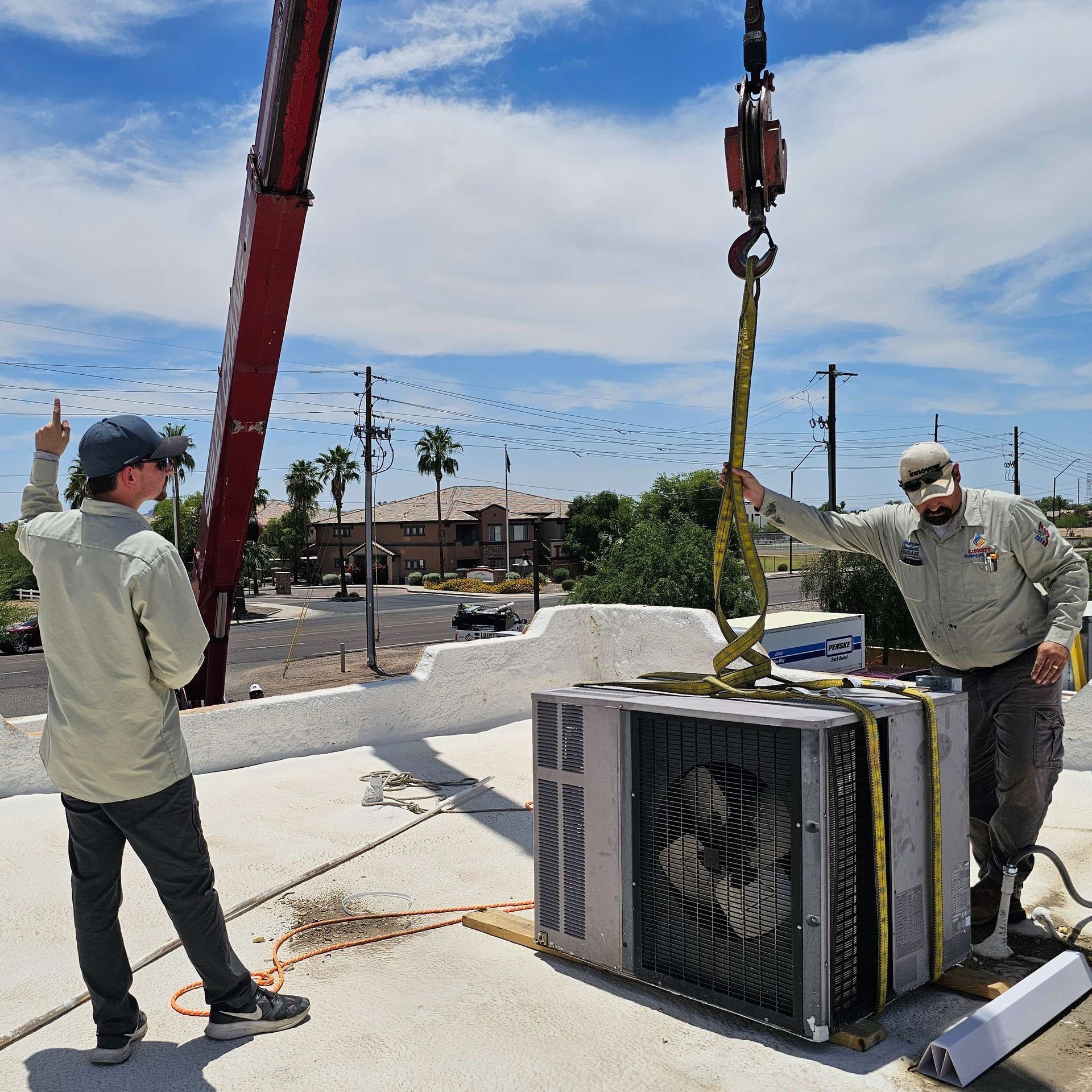 Two men using a crane to lift an AC unit onto a building roof under a blue sky.