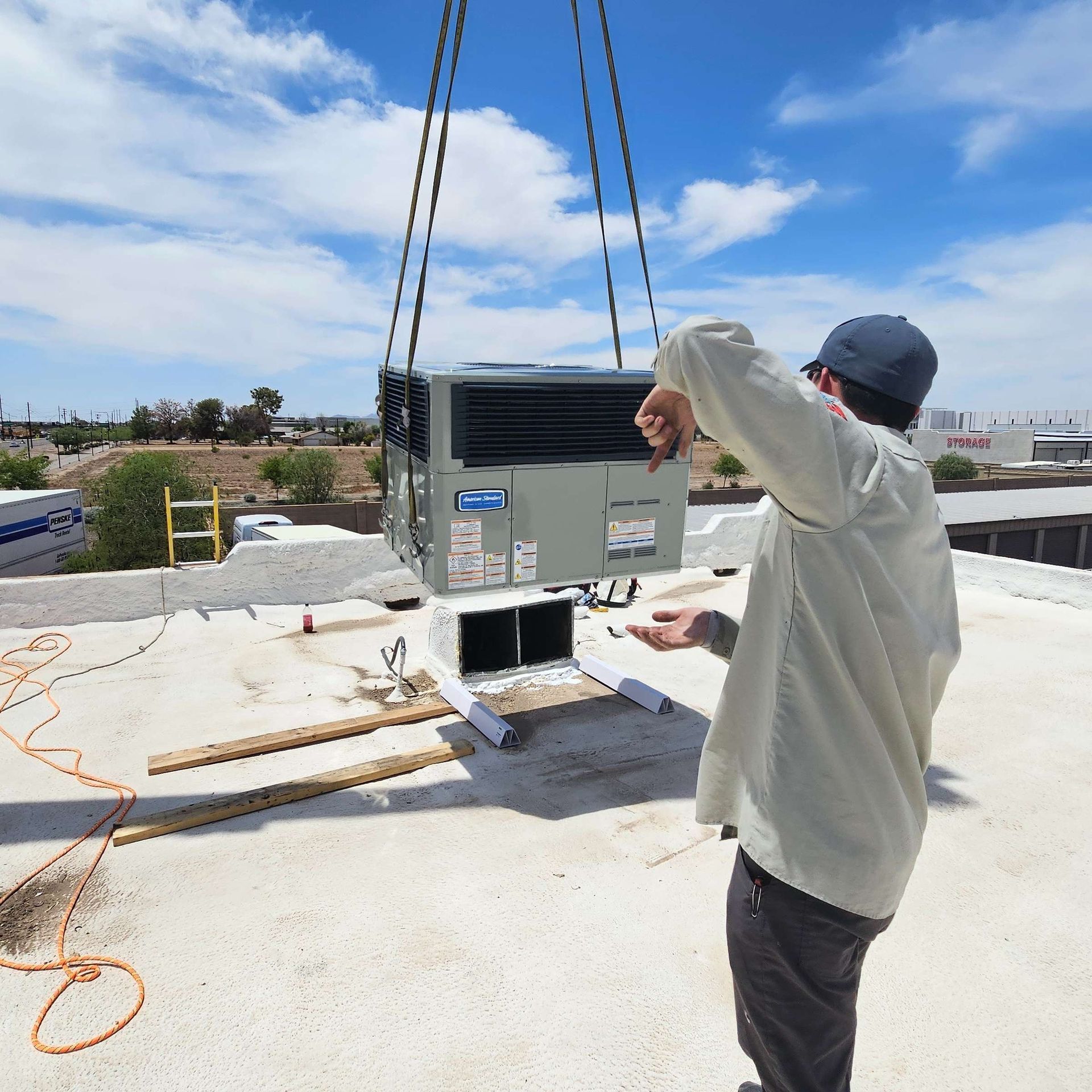 Person guides an AC unit being lifted by a crane onto a rooftop on a sunny day.