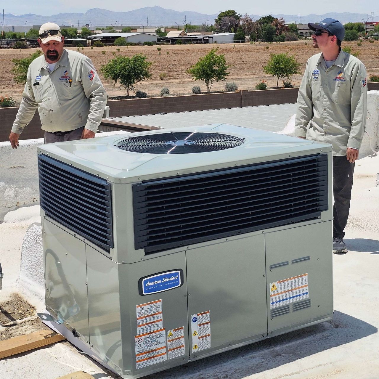 Two men lifting an air conditioning unit on a rooftop. The unit is silver, and the sky is blue.