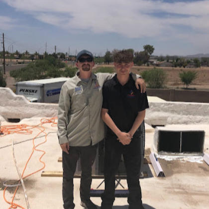 Two men pose on a rooftop. One has an arm around the other's shoulder. A truck is in the background.