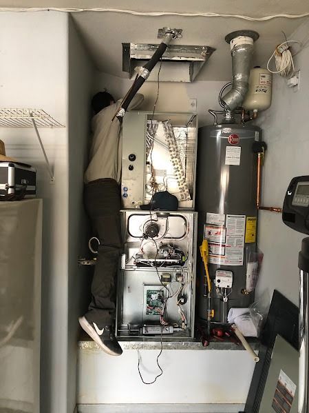 Person in a utility room working on a furnace and water heater. Gray and white appliances in a small space.