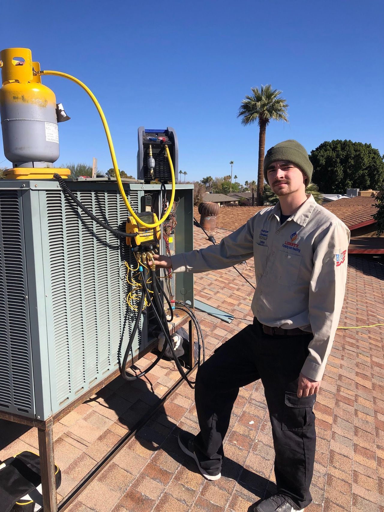 HVAC technician on a rooftop points to AC unit being refilled with yellow tank nearby. Bright sky.