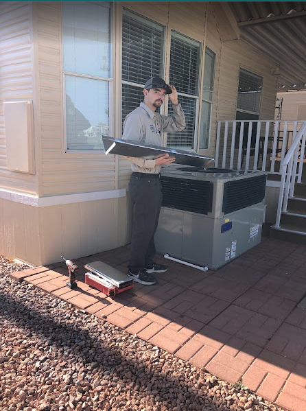 HVAC technician next to an AC unit, holding parts outdoors; wearing a hat and work uniform.