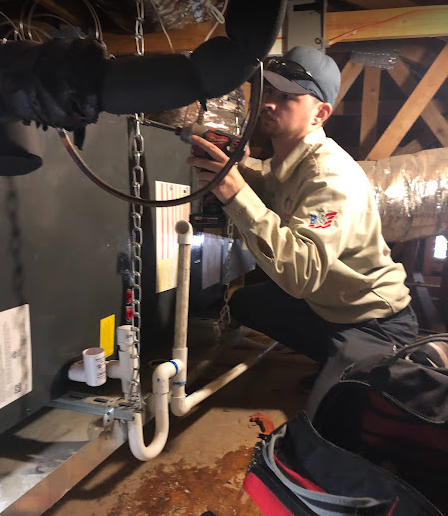 HVAC technician kneels, working on system in a crawlspace, wearing a hat and uniform.