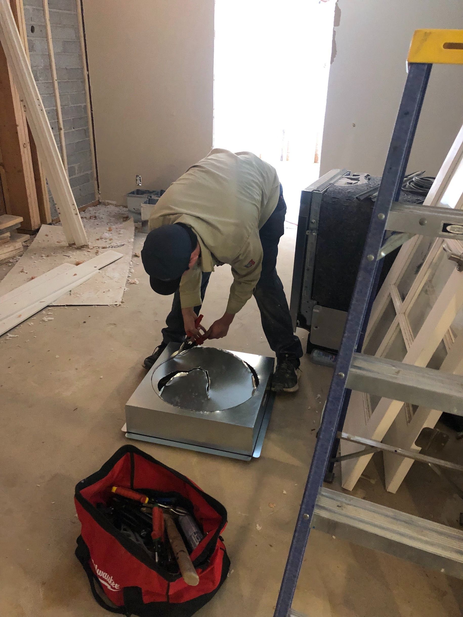 A worker in a construction zone is working with metal vent components. Red tool bag nearby.