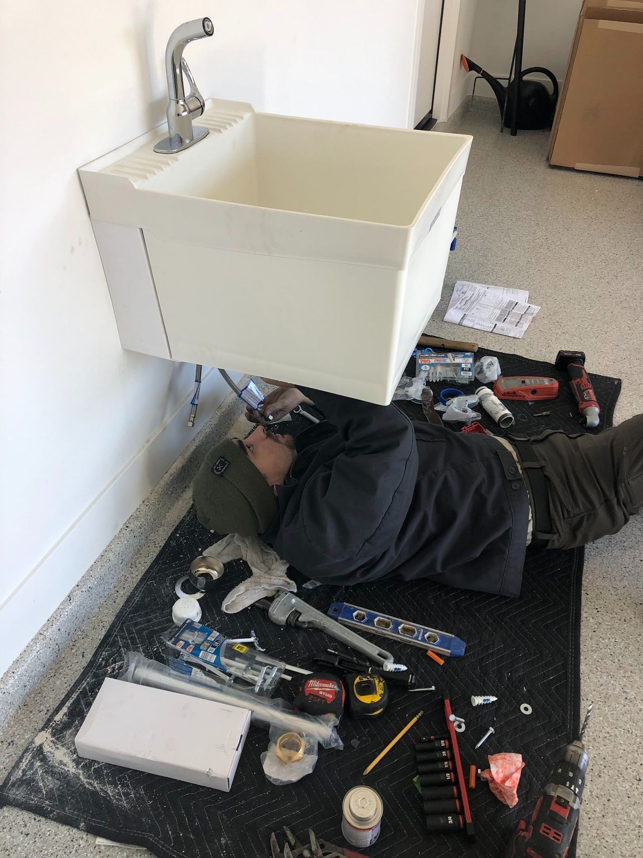 Person installing a utility sink, lying on a mat with tools scattered around in a garage.