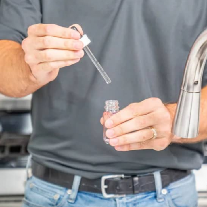Person holding dropper over small bottle near a faucet, likely testing water quality.