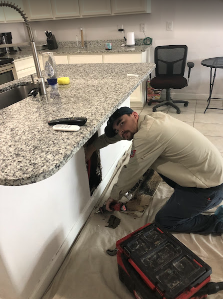 Man kneels, working under a kitchen island. White cabinets, granite countertop. Tools and red toolbox are visible.