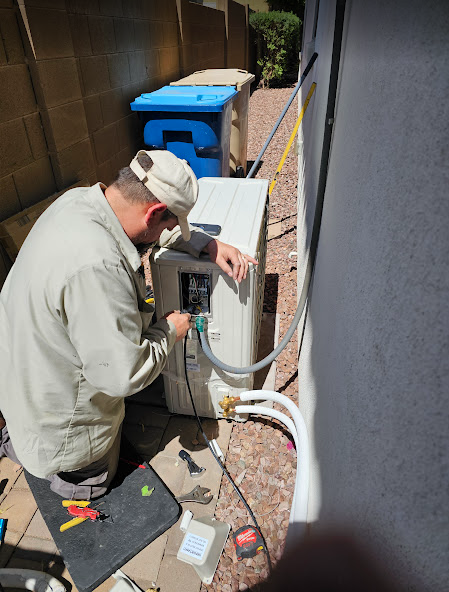Man in beige uniform working on outdoor air conditioning unit next to a wall; trash bins nearby.