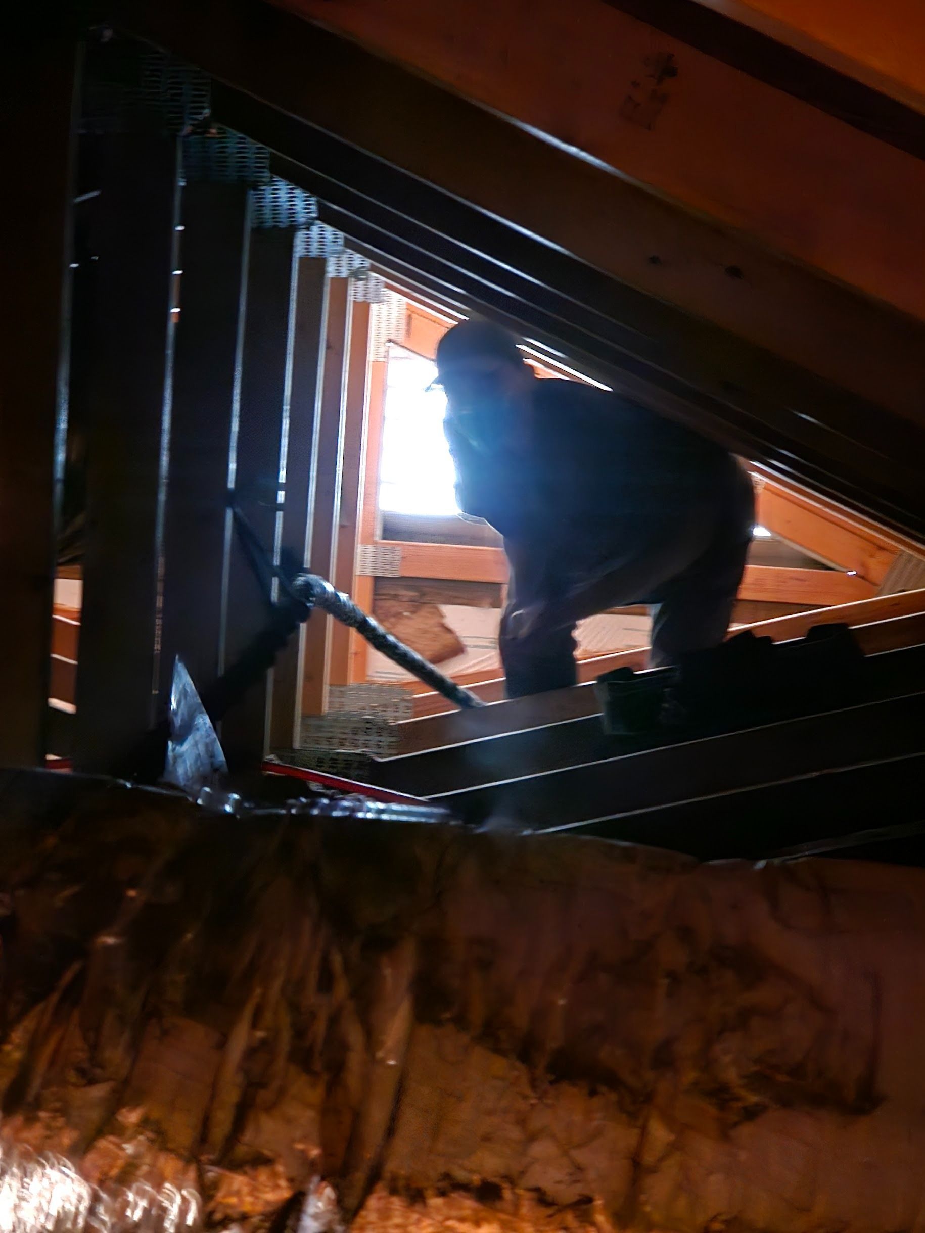 Man in an attic, likely working on insulation. Dim lighting, wood beams, and a small opening visible.