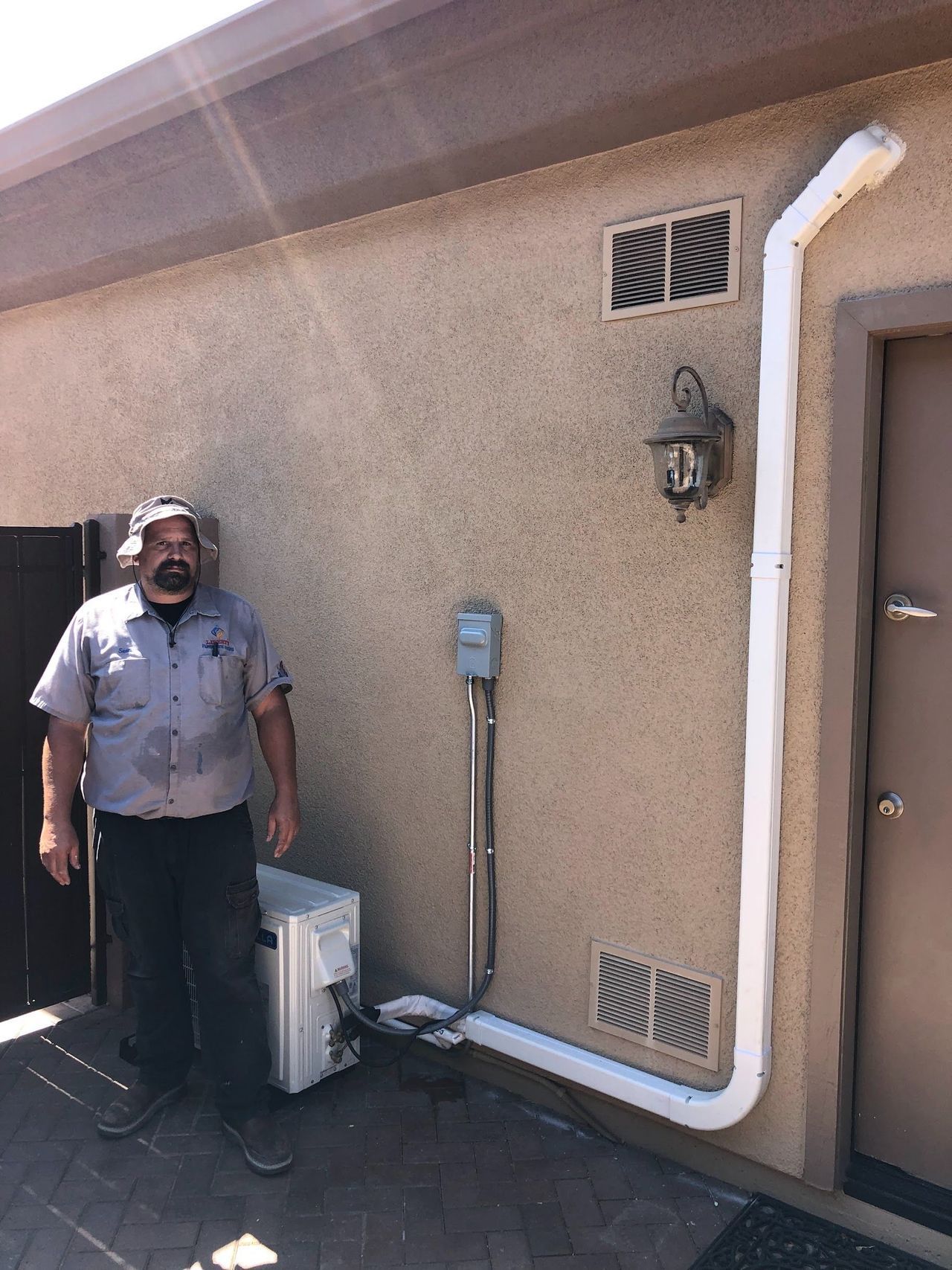 A man stands beside an AC unit. A white pipe runs up the wall. The wall is beige.