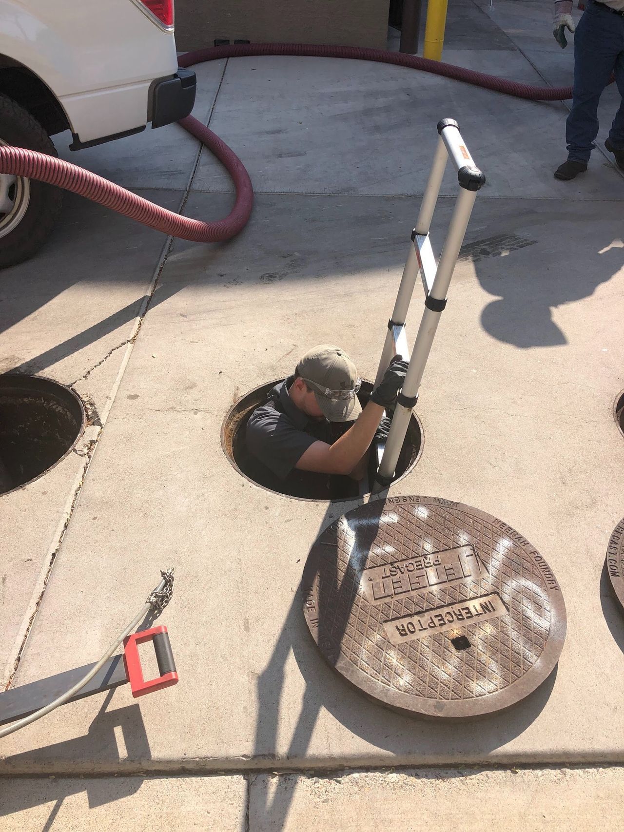 Man in manhole with ladder; next to a sewer cover. Large hose connected to a truck.