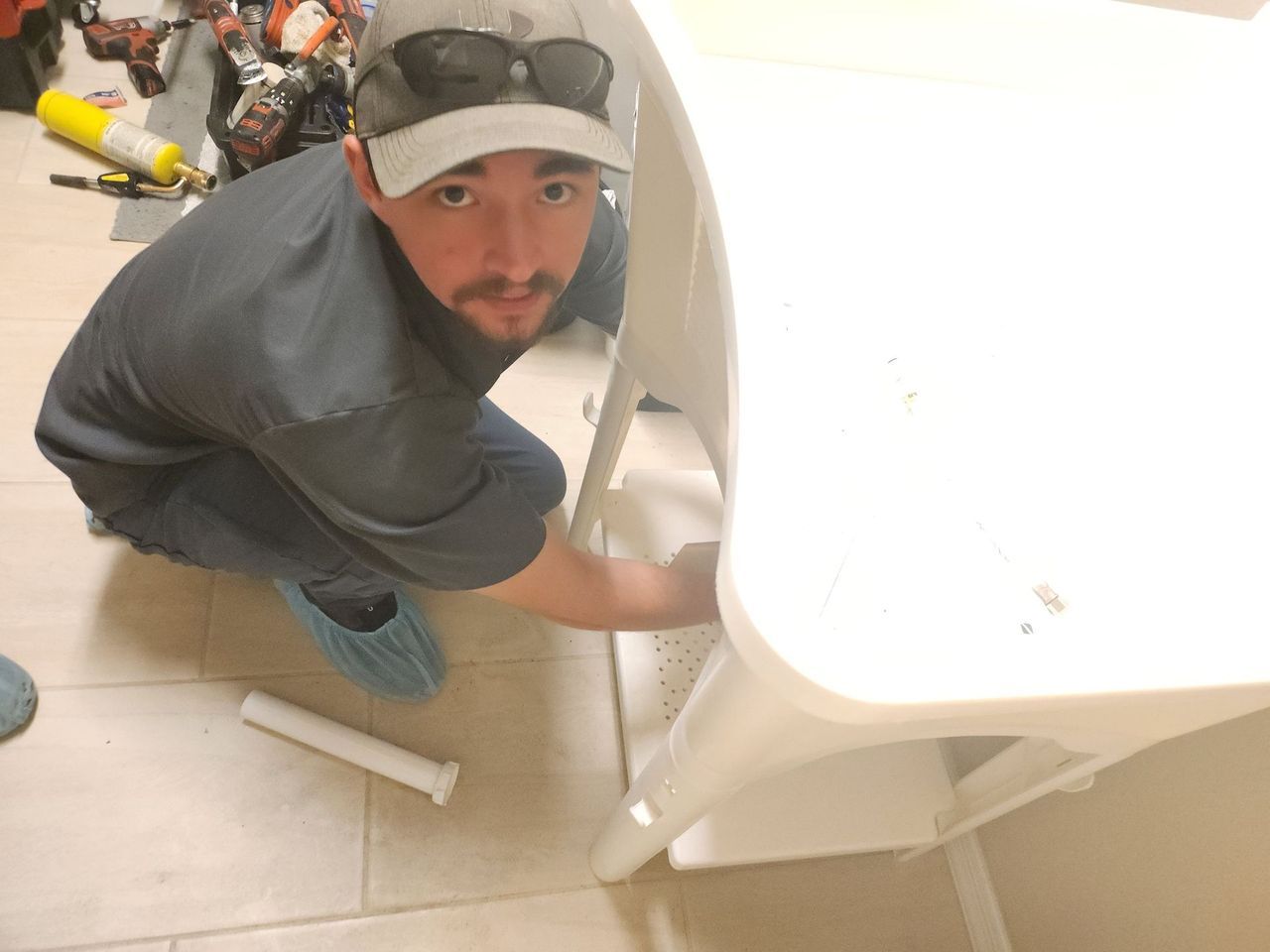 Man in gray shirt and hat repairs a white washing machine, squatting.