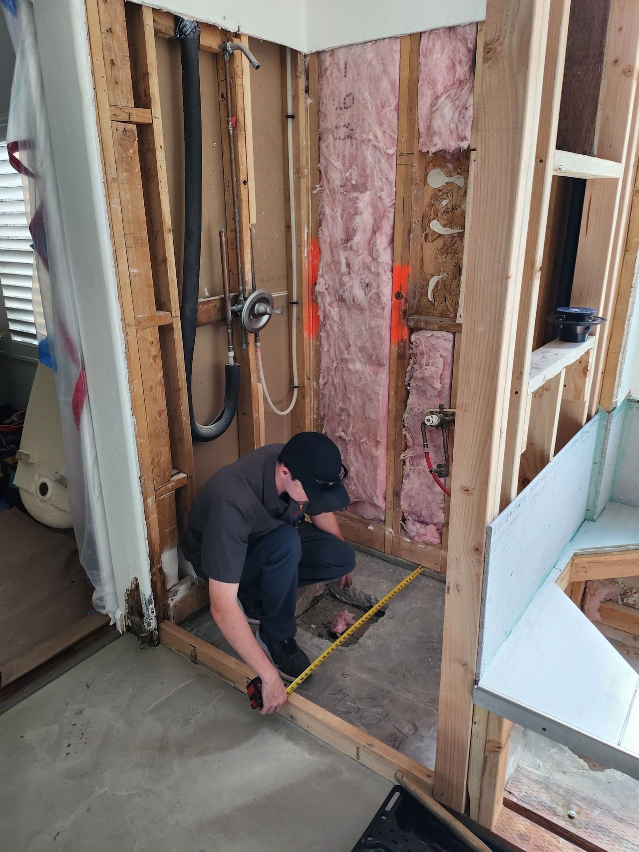 Man measures inside a bathroom under construction, framed by wooden studs, insulation visible.