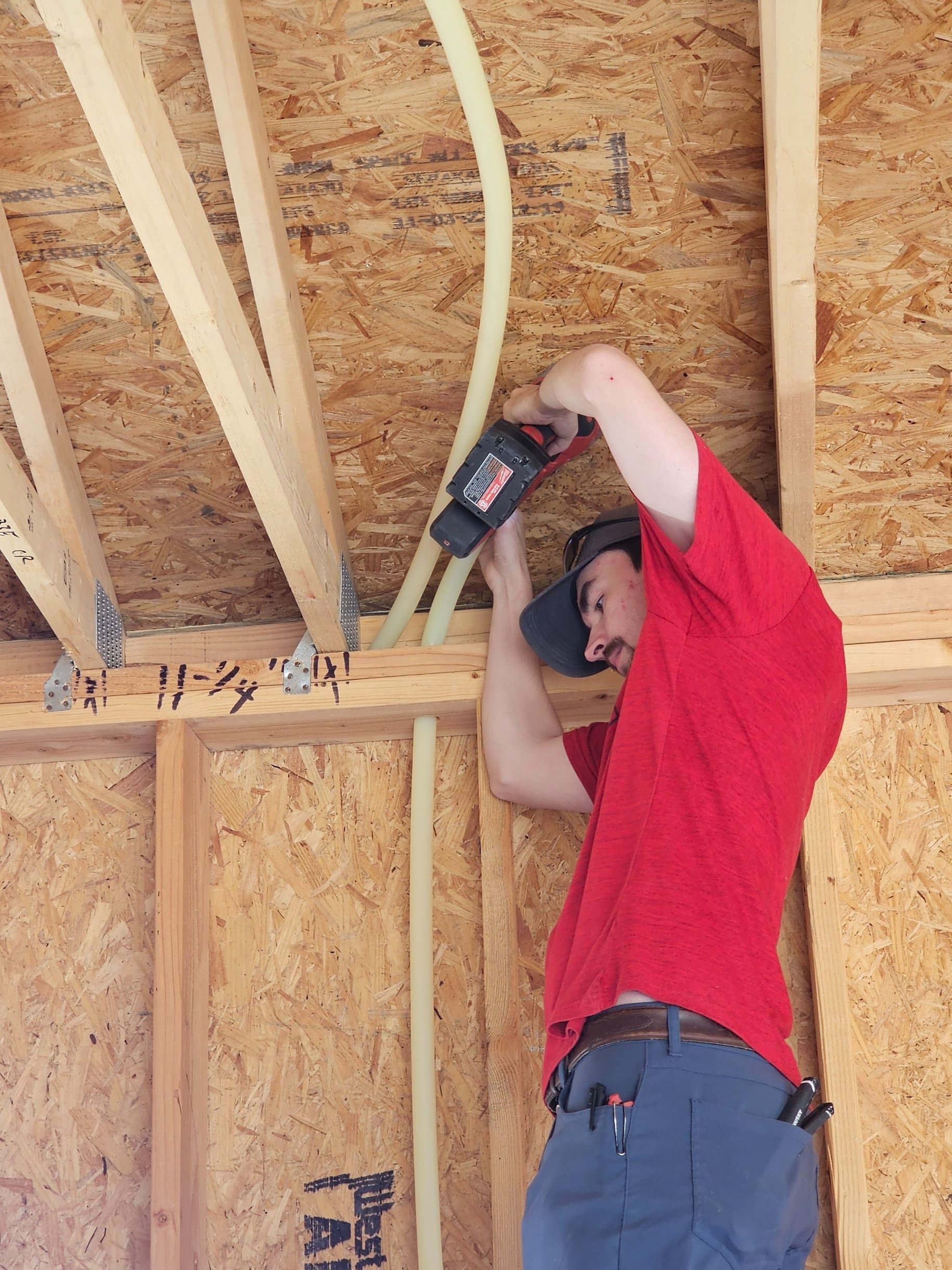 Man in red shirt using a drill on yellow tubing in wooden wall. Construction setting.