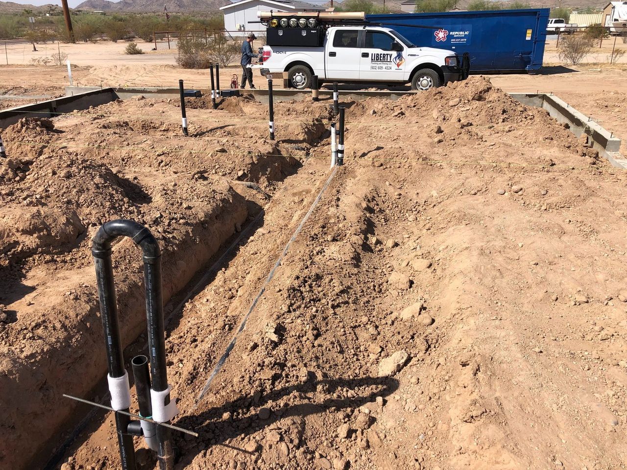 Trenches with plumbing pipes at a construction site, worker nearby, truck in background.