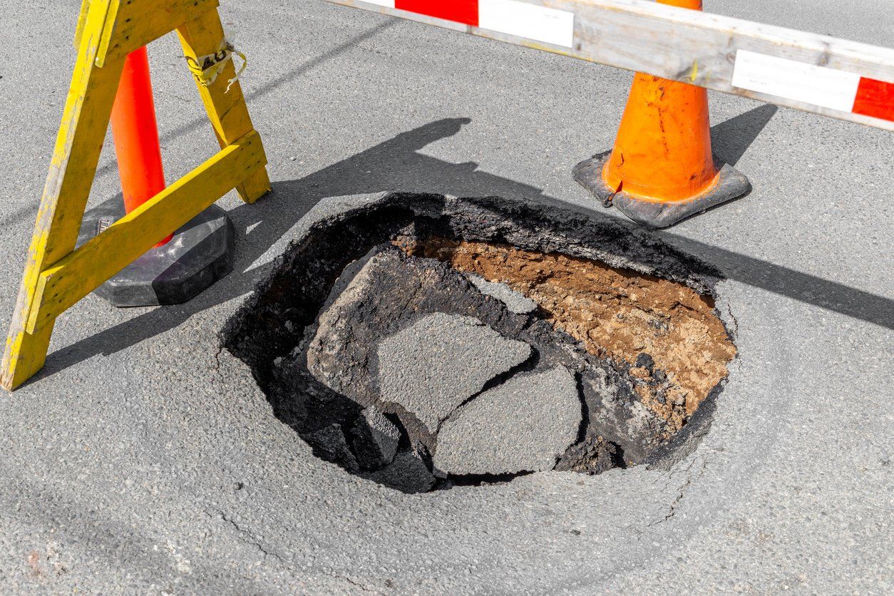 large pothole needing asphalt repairs surrounded by a yellow barrier and an orange traffic cone