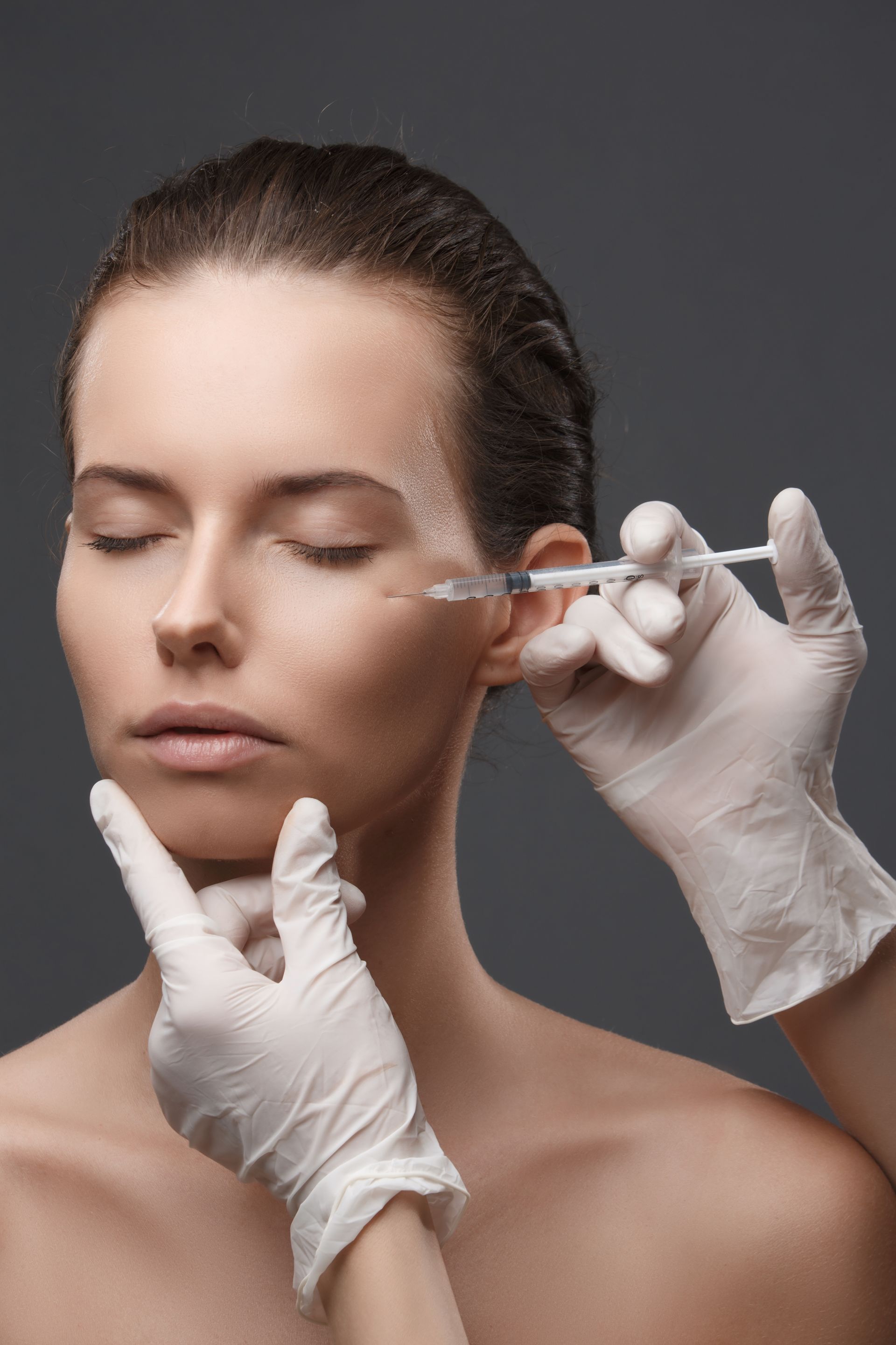 Woman receiving cosmetic injection near eye, wearing white gloves, against gray background.
