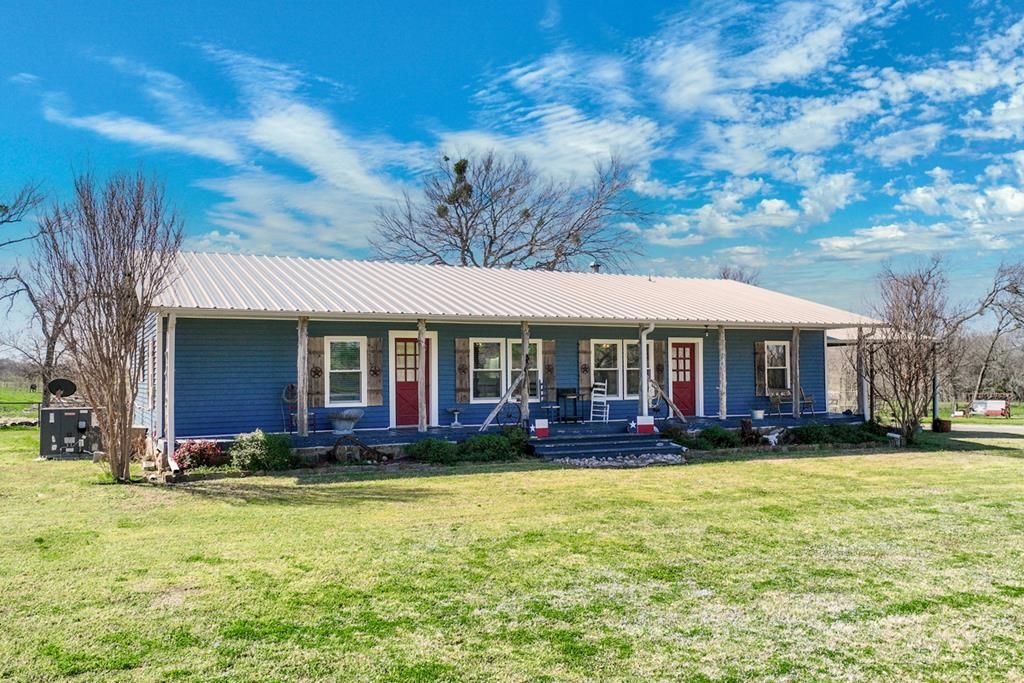 Blue ranch house with red doors, metal roof, and green yard under a blue sky.