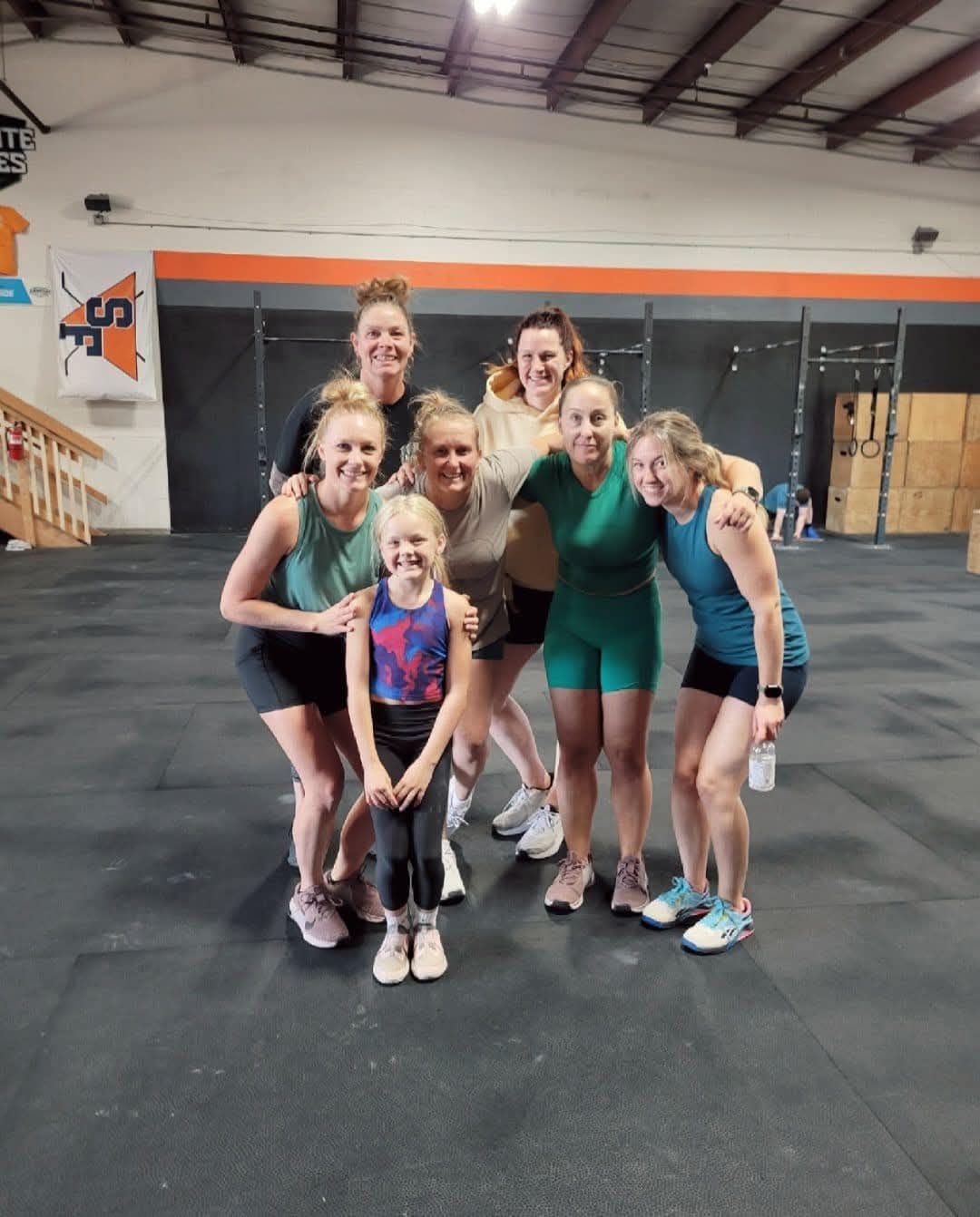 Group of women and a child in a gym, smiling, posing for a photo. Athletic wear, dark floor, and colorful wall.