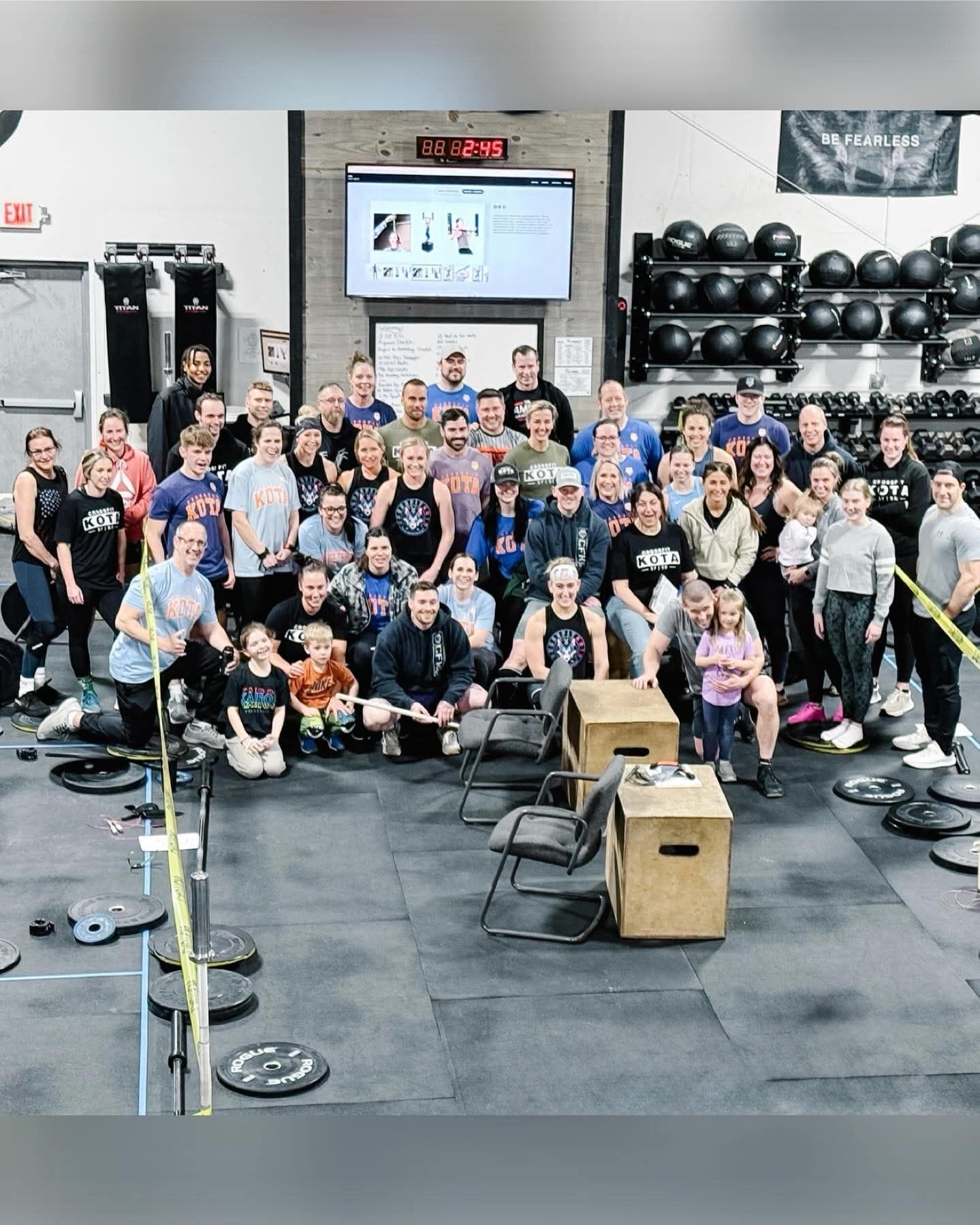 Group of people at a gym, posing for a photo. Some seated, some standing. Fitness equipment visible.