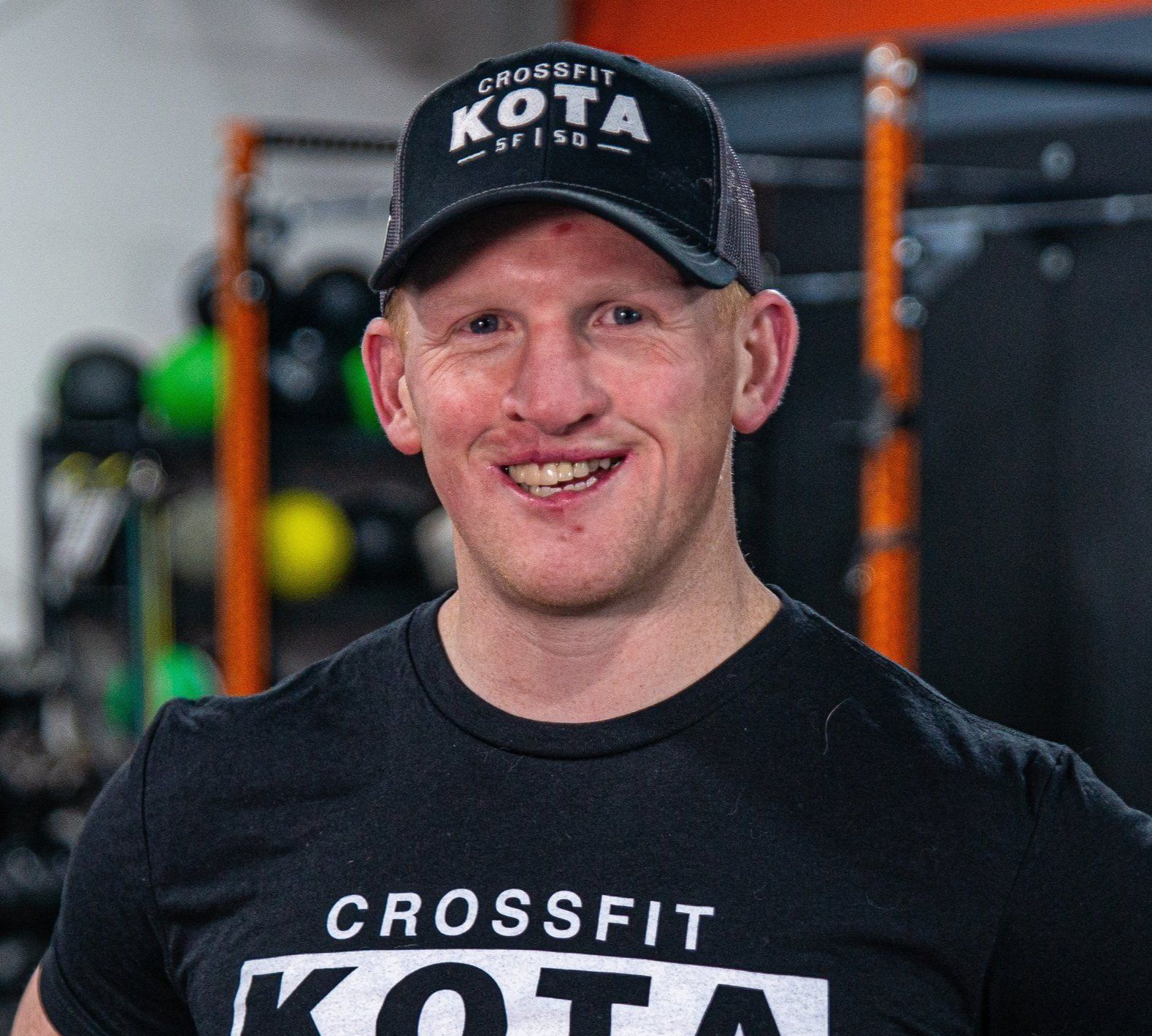 Man wearing a CrossFit KOTA cap and t-shirt, smiling, in a gym setting.