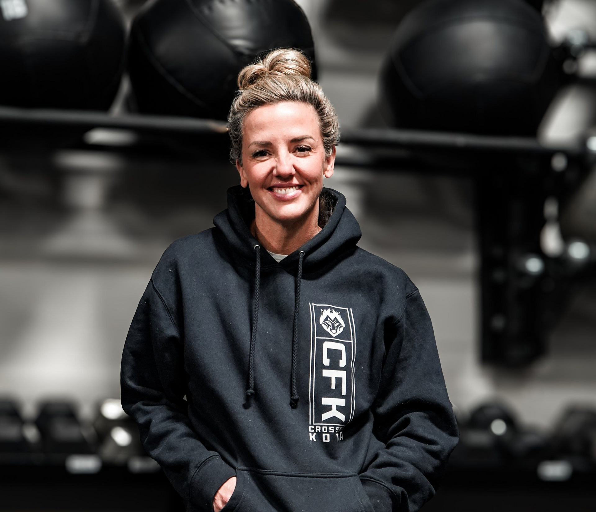 Woman in a black hoodie and leggings smiles in a gym, leaning on dumbbells.