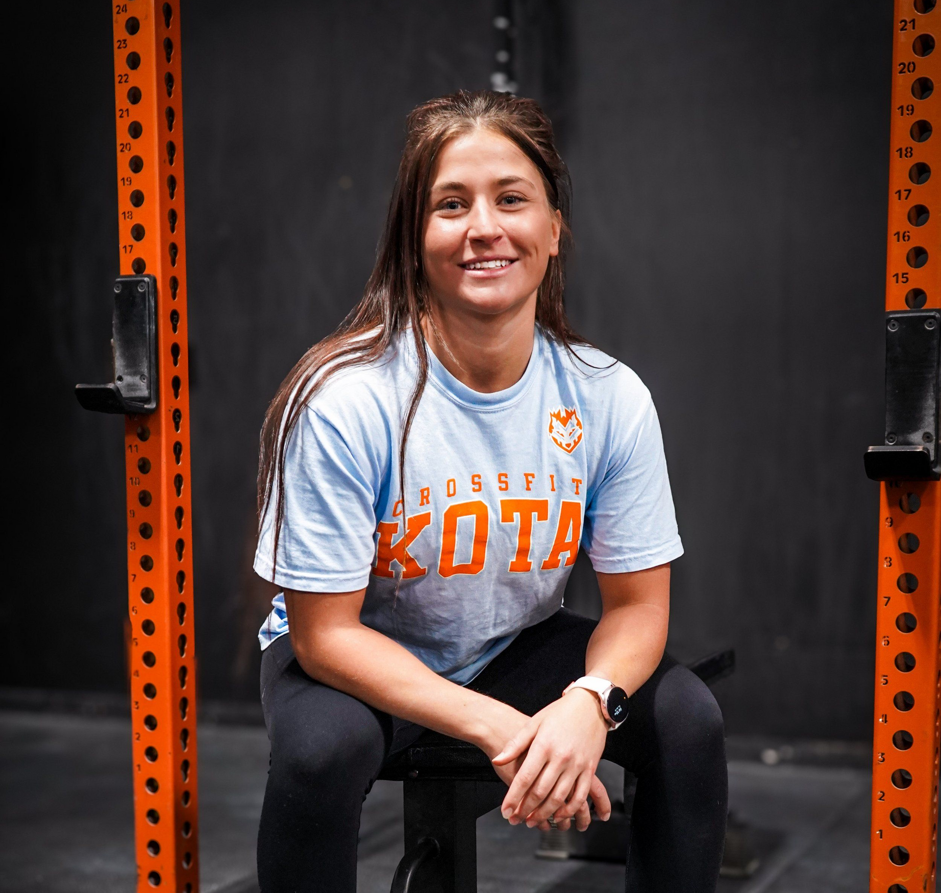 Woman in light blue t-shirt with orange writing, seated, smiling, next to an orange weight rack, in a gym.