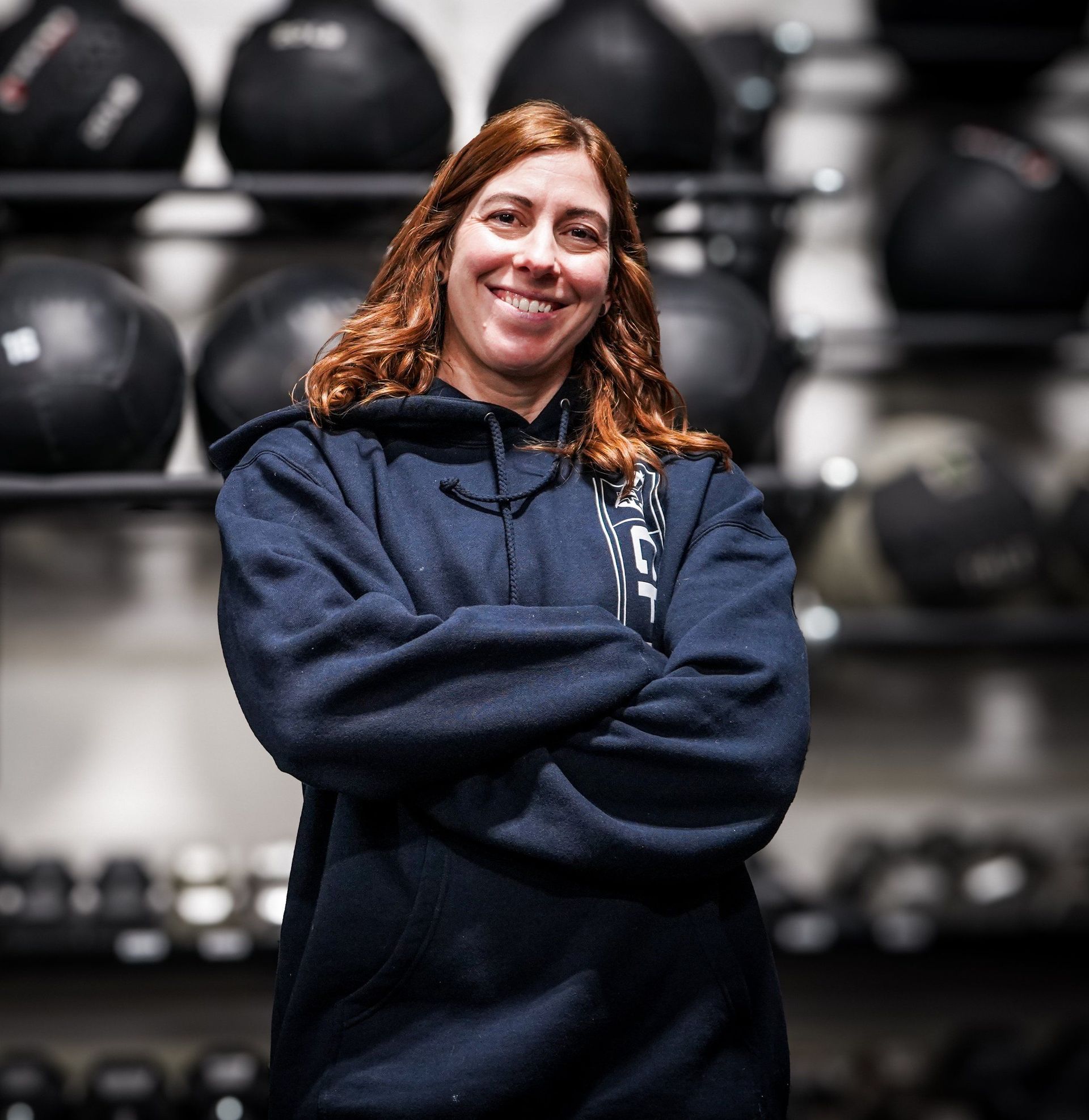 Woman with crossed arms smiling in front of weight training equipment; she wears a blue hooded sweatshirt.