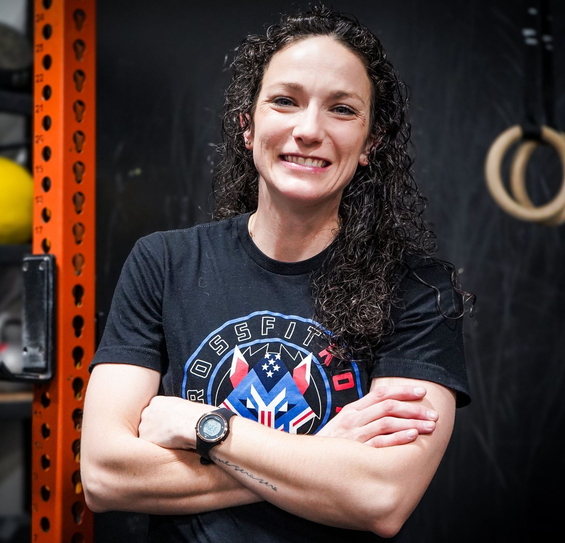Woman with crossed arms, smiling, wearing a black shirt in a gym.