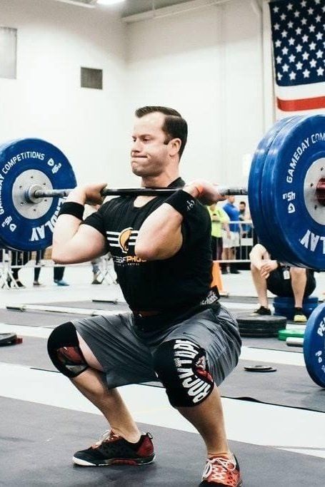 Weightlifter in a deep squat, barbell overhead, at a competition. He wears black shirt, grey shorts, and knee sleeves.