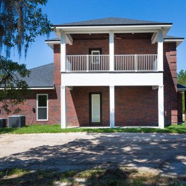 Two-story brick house with white columns, balcony, and dark roof on a sunny day.