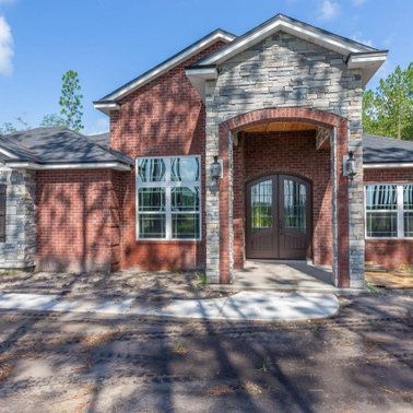Brick house with stone accents, arched entryway, double doors, and multiple windows under a blue sky.