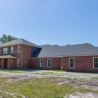 Brick two-story house with dark roof and multiple windows in a grassy field under a blue sky.