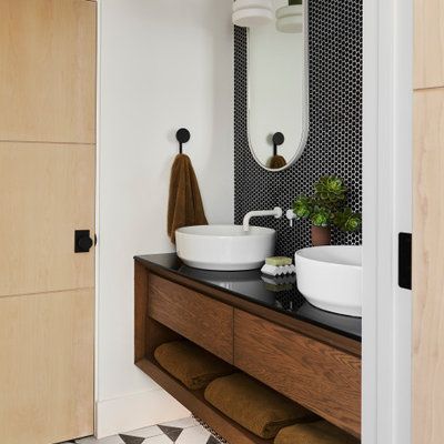 Bathroom with floating wooden vanity, white vessel sinks, black tile wall, and oval mirror.
