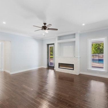 Empty living room with dark wood floor, fireplace, and large windows overlooking a pool.