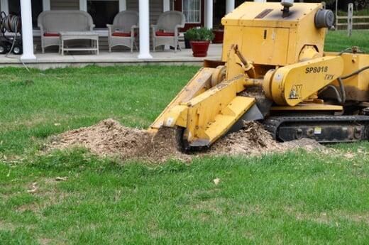 A yellow tractor is cutting a tree stump in a yard.