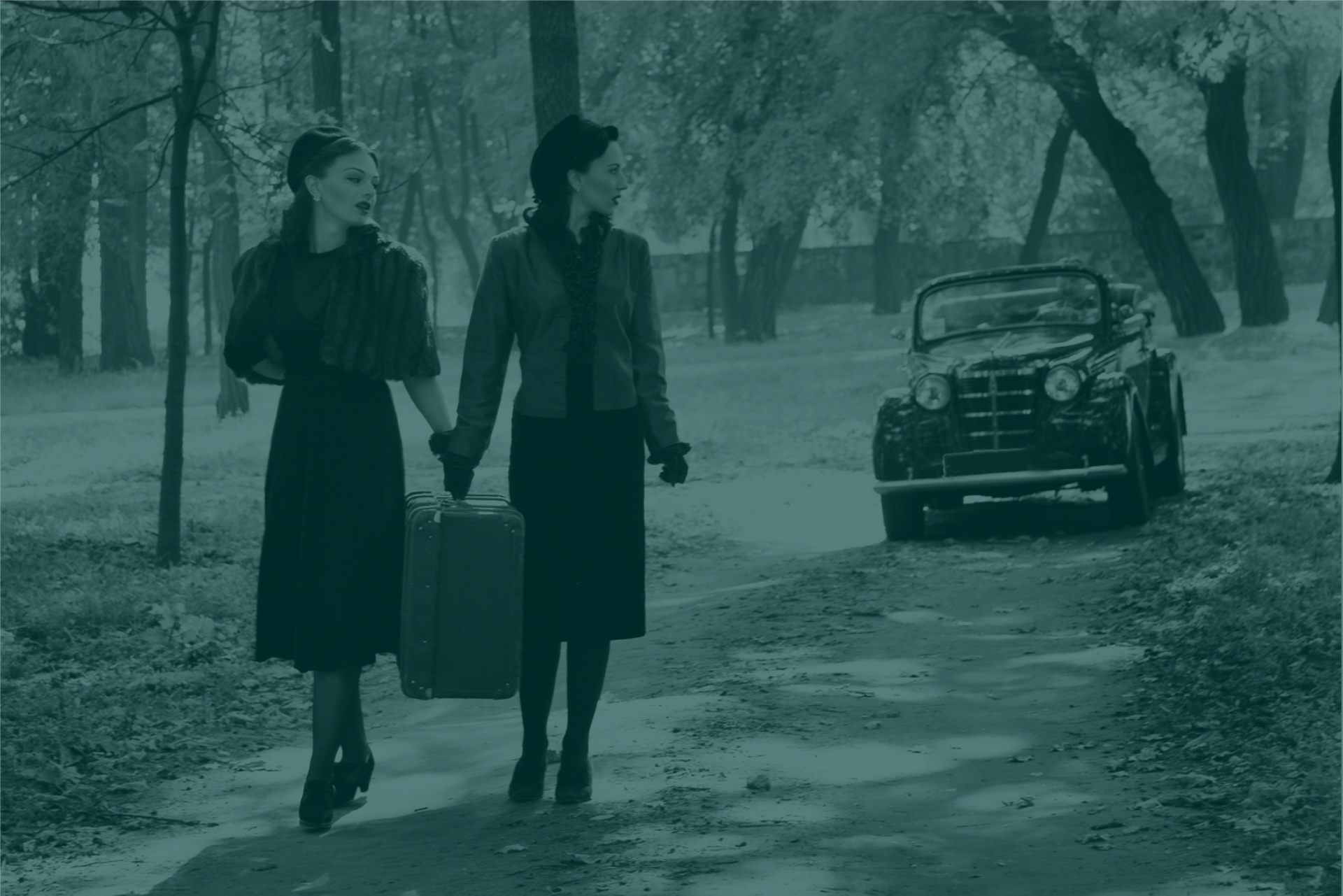 Duas mulheres de vestido preto em uma estrada de terra carregando uma mala com um automóvel ao fundo