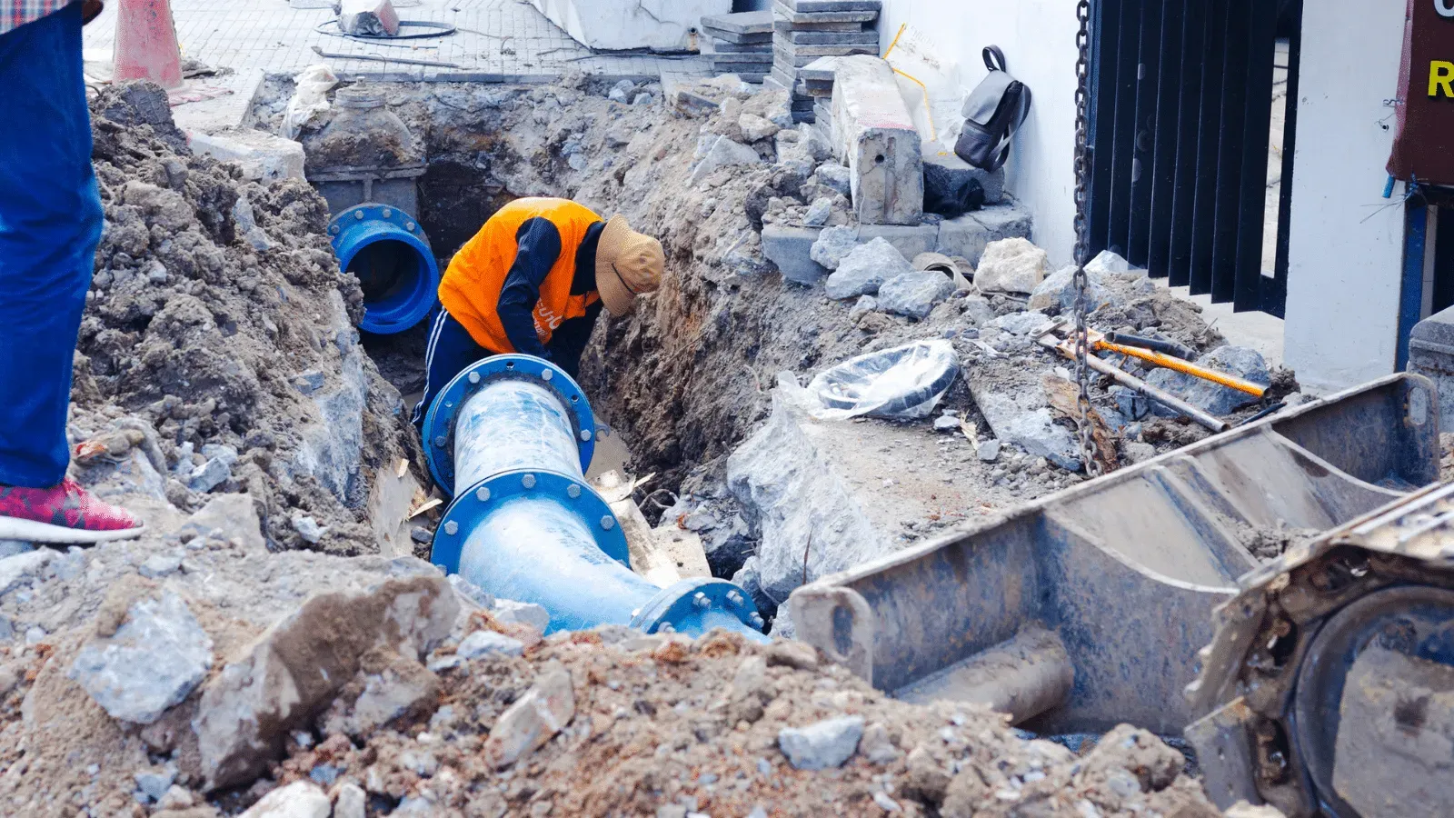 Workers in an orange vest installing large blue pipes in a trench, surrounded by debris.