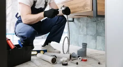 Plumber kneeling under a cabinet, working on pipes with a wrench and tools.