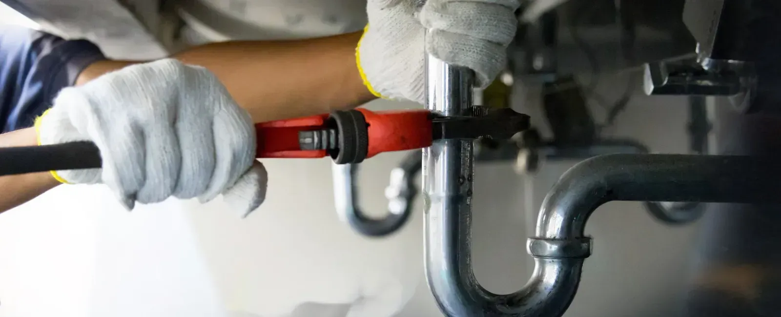 Person wearing gloves uses a red wrench on plumbing pipes.